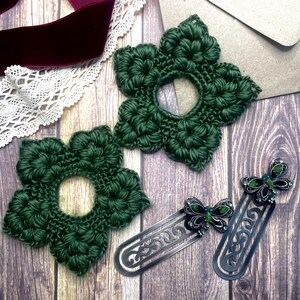 May include: Two dark green crocheted flower-shaped coasters and two silver metal butterfly bookmarks with green accents are displayed on a wooden surface. A burgundy velvet ribbon and white lace trim are also visible.