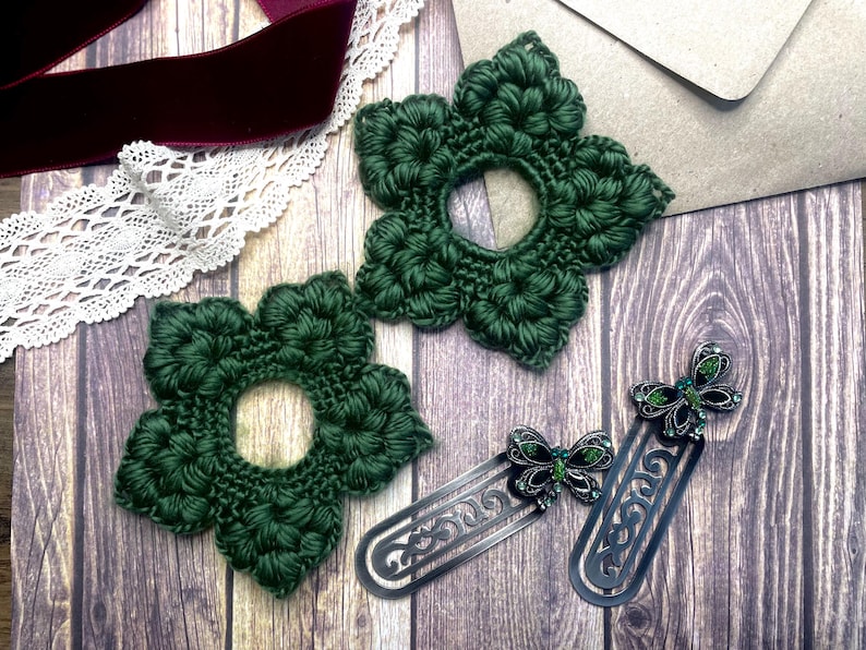 May include: Two dark green crocheted flower-shaped coasters and two silver metal butterfly bookmarks with green accents are displayed on a wooden surface. A burgundy velvet ribbon and white lace trim are also visible.