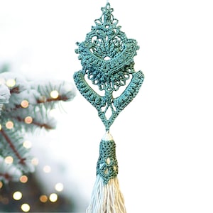 Może przedstawiać: Turkusowa, szydełkowa ozdoba z chwostem. Ozdoba ma szczegółowy, ażurowy wzór i kształt łzy. Chwost wykonany jest z kremowej przędzy. Tło jest rozmyte, z ośnieżoną gałęzią drzewa iglastego.
