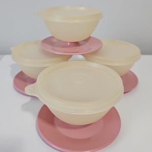 May include: A set of four small, round, beige plastic bowls with matching translucent lids, each resting on a solid pink circular saucer. The bowls are arranged in a tiered formation, with one bowl positioned in the front and three behind it.