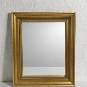 Puede incluir: Un espejo rectangular dorado con un marco grueso y ornamentado. El marco tiene un diseño texturizado y acanalado y un cálido acabado dorado antiguo. El espejo refleja un fondo neutro de color claro.