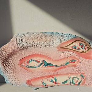 Könnte beinhalten: Ein handgefertigtes Keramikkunstwerk mit strukturierter Oberfläche. Das Kunstwerk zeigt eine Palette aus Rosa, Blau und Gold mit abstrakten Formen und Mustern. Das Stück hat eine einzigartige, organische Form und ist wahrscheinlich ein dekorativer Gegenstand.