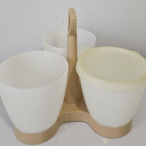 Puede incluir: Un servidor de condimentos de plástico beige con tres tazas blancas translúcidas sostenidas por un asa central. Una taza está cubierta con una tapa beige a juego, mientras que las otras dos tazas permanecen abiertas y sin tapa.