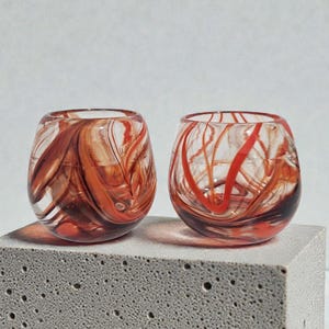 May include: Two clear, stemless glasses with swirling red and brown patterns. The glasses are on a grey, textured surface. The glass design features a unique, artistic style.
