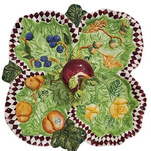 Puede incluir: Un plato de servicio de cerámica de cuatro secciones con un asa central en forma de manzana roja. El plato presenta un patrón de hojas verdes con moras, bellotas, melocotones y rosas amarillas en relieve. Borde con patrón de cuadros rojo y blanco.