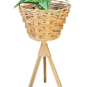 Puede incluir: Un soporte para plantas de mimbre tejido con una base de tres patas. La maceta estilo cesta contiene una planta verde. El soporte está hecho de madera clara y mimbre, perfecto para la decoración interior o exterior.