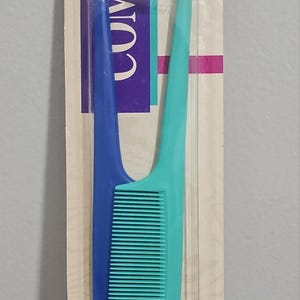 May include: A dual-sided hair comb in a clear plastic package. One side is blue, the other is teal. The comb has fine teeth for styling and detangling hair. The packaging has the word "VOLUME" printed on it.