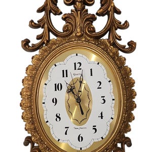May include: Ornate gold-colored wall clock with a decorative frame. The clock face is white with black numerals and hands. The clock reads "New Haven" and "Quartz". The frame features intricate scrollwork.