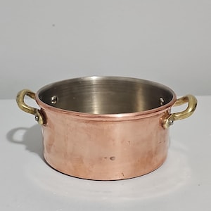 May include: A small, copper-colored cooking pot with two brass handles. The pot has a shiny, metallic interior and a rounded shape. It is designed for cooking and serving small portions of food.