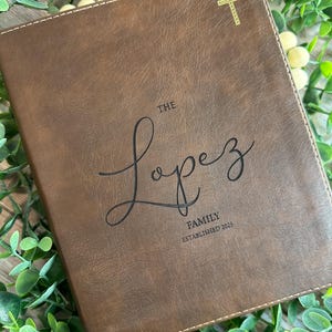 May include: A brown leather journal with the name "Lopez" in cursive script. The text "FAMILY ESTABLISHED 2025" is below the name. A gold cross is in the upper right corner. The journal is surrounded by green leaves.