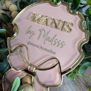 May include: A pink and gold decorative plaque with the text "MANIS by Madsss" in gold lettering. The plaque has a bow design at the bottom and is surrounded by greenery.
