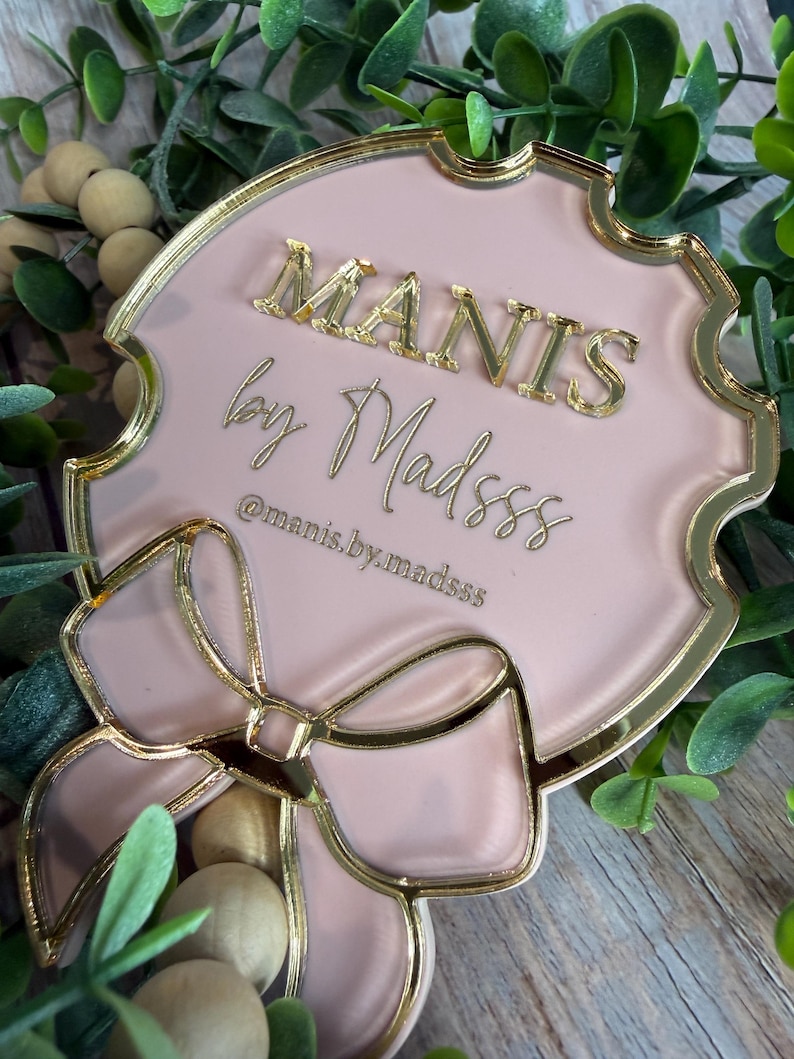 May include: A pink and gold decorative plaque with the text "MANIS by Madsss" in gold lettering. The plaque has a bow design at the bottom and is surrounded by greenery.