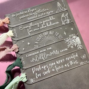 Peut inclure: Marque-pages en acrylique transparent avec des citations inspirantes et des éléments décoratifs. Chaque marque-page présente une phrase différente en lettres blanches, comme "Soyez joyeux dans l'espoir". Des rubans de couleurs variées sont attachés.