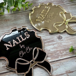 May include: Two decorative nail art signs. One is gold-toned, the other is black with gold trim. Both signs have the word "NAILS" in raised lettering, along with a bow design. The black sign also includes the text "by Chu Chi".