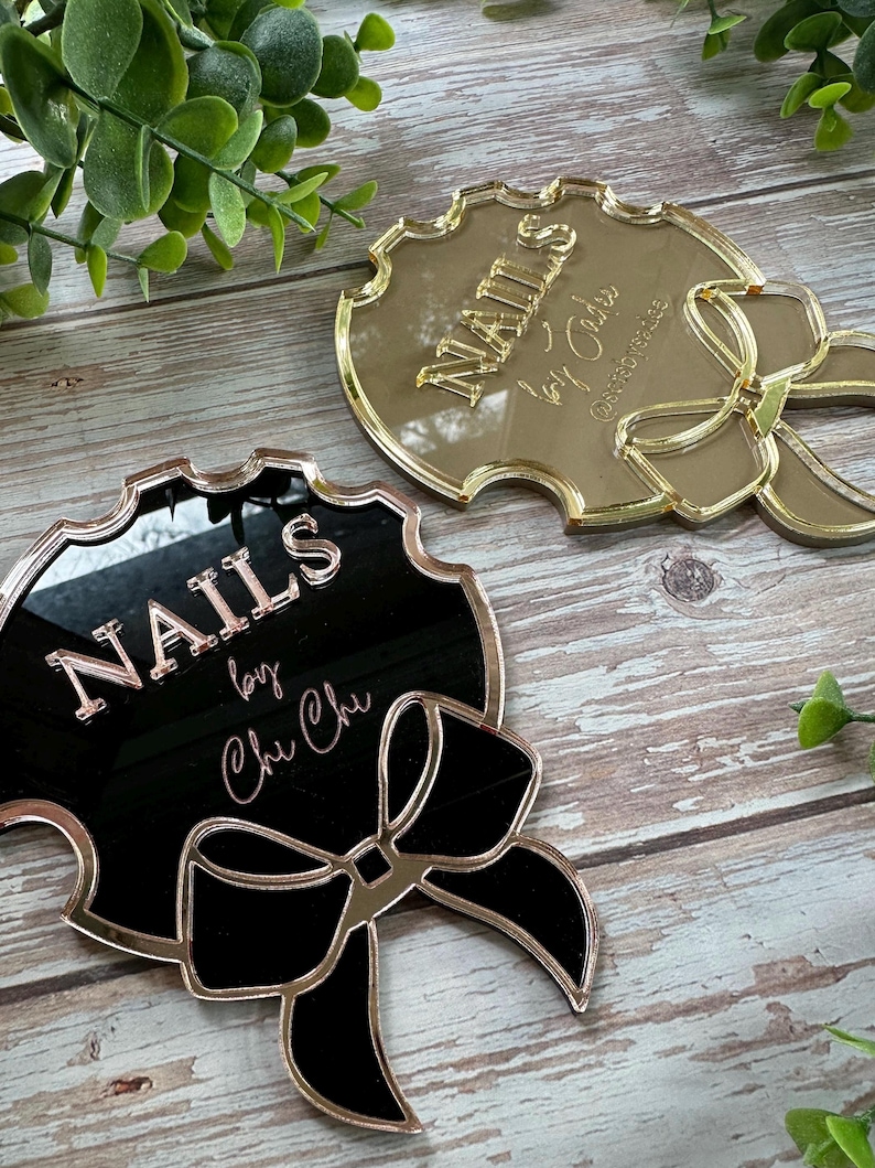 May include: Two decorative nail art signs. One is gold-toned, the other is black with gold trim. Both signs have the word "NAILS" in raised lettering, along with a bow design. The black sign also includes the text "by Chu Chi".