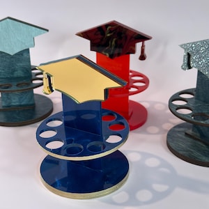 Puede incluir: Tres porta lápices con forma de gorro de graduación, uno azul, uno rojo y uno verde. Cada porta lápices tiene una base redonda con varios agujeros para lápices. Los gorros están decorados con diferentes patrones.