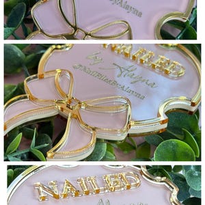 May include: A pink and gold decorative sign with the text "NAILED by Alayna" and the Instagram handle "@NailFilesByAlayna". The sign has a bow design and a gold border.