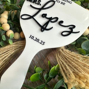 May include: A white, paddle-shaped wedding sign with black lettering that reads "MRS. Lopez" and "PREPARING TO BE" with the date "10.20.25". The sign is surrounded by greenery and twine.
