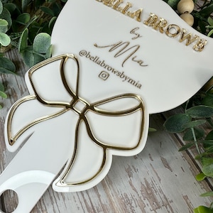 Op de afbeelding: Een witte handspiegel met een goudkleurige strik aan de basis. Het oppervlak toont de tekst BELLA BROWS by Mia en de Instagram-gebruikersnaam @bellabrowsbymia in gouden letters. De spiegel heeft een rond gat in het handvat.