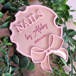 May include: A pink sign with the word "NAILS" in raised letters, the name "Abby" in cursive, and the Instagram handle "@abs.nails_". The sign has a bow design and is surrounded by green foliage.
