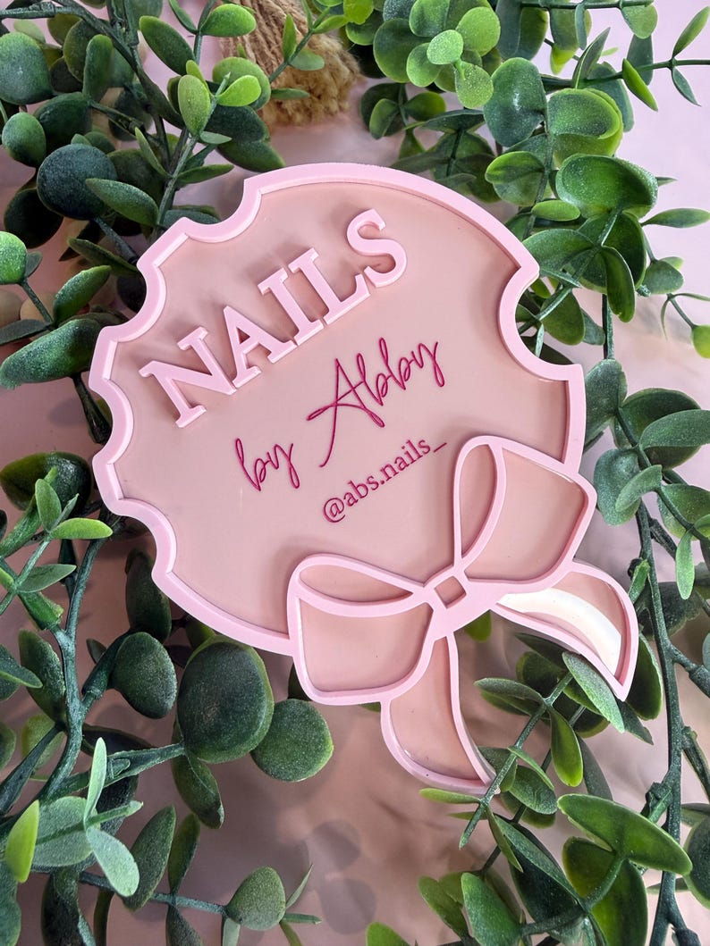 May include: A pink sign with the word "NAILS" in raised letters, the name "Abby" in cursive, and the Instagram handle "@abs.nails_". The sign has a bow design and is surrounded by green foliage.