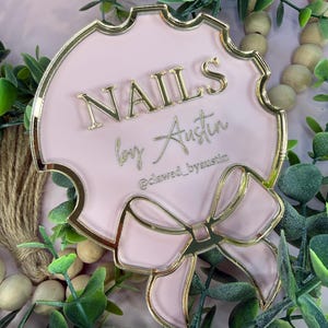 May include: A pink and gold sign with the words "NAILS by Austin" in gold lettering. The sign has a decorative bow at the bottom and a scalloped edge. The Instagram handle "@clawed_byaustin" is also visible.