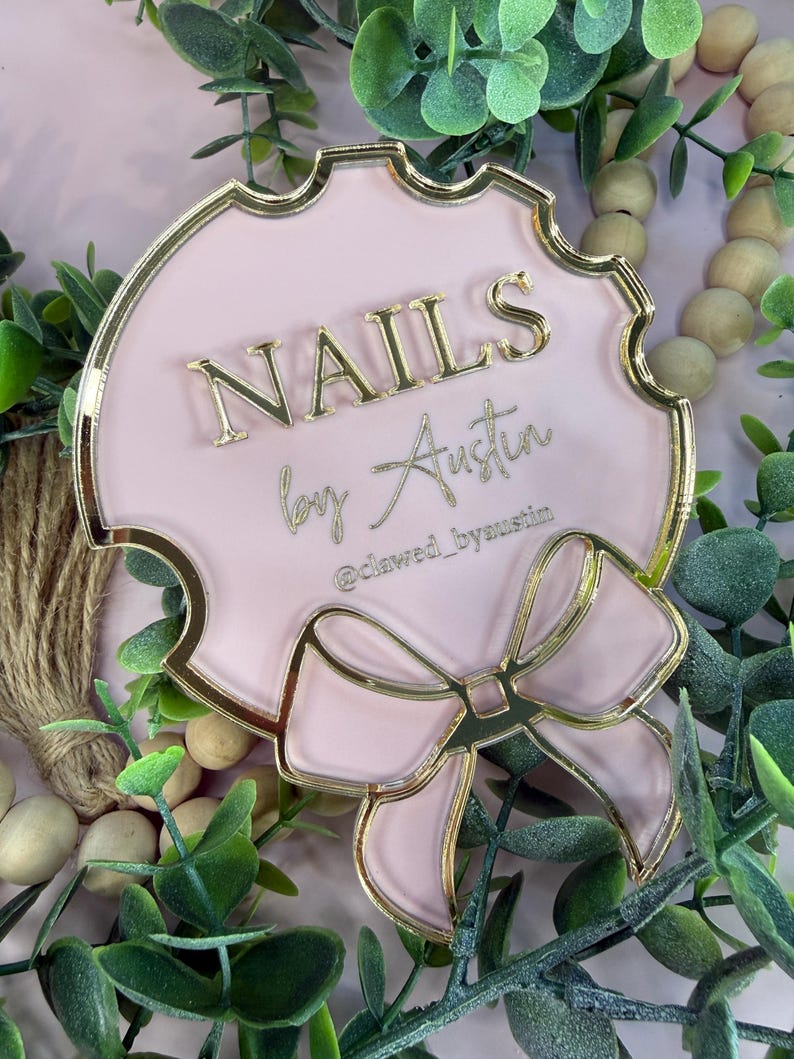 May include: A pink and gold sign with the words "NAILS by Austin" in gold lettering. The sign has a decorative bow at the bottom and a scalloped edge. The Instagram handle "@clawed_byaustin" is also visible.