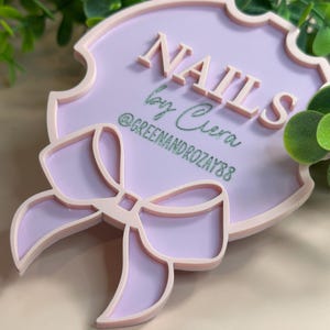 May include: A light purple sign with the word "NAILS" in raised letters. The sign also includes the text "by Ciera" and the Instagram handle "@GREENANDROZAY88". A bow design is at the bottom of the sign.