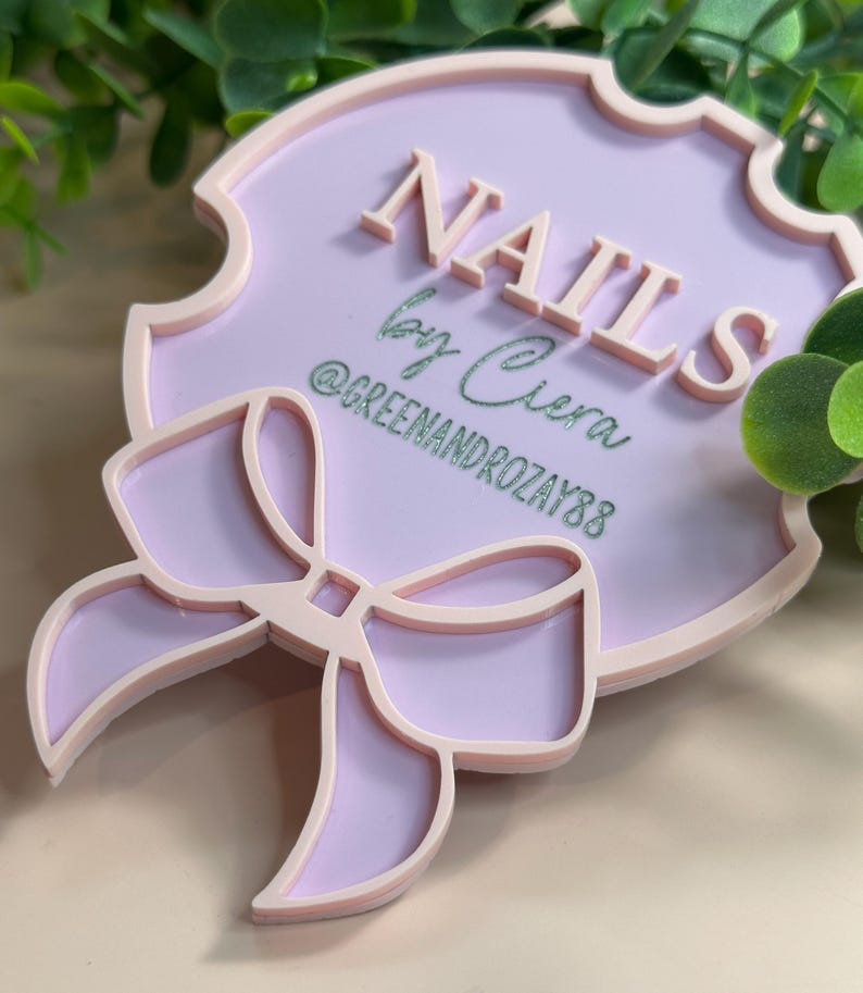 May include: A light purple sign with the word "NAILS" in raised letters. The sign also includes the text "by Ciera" and the Instagram handle "@GREENANDROZAY88". A bow design is at the bottom of the sign.