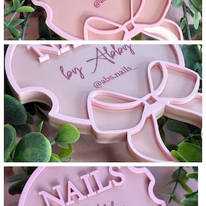 May include: A pink, layered sign with the text "NAILS by Abby" and the Instagram handle "@abs.nails_". The sign has a bow design at the bottom and is surrounded by green foliage.