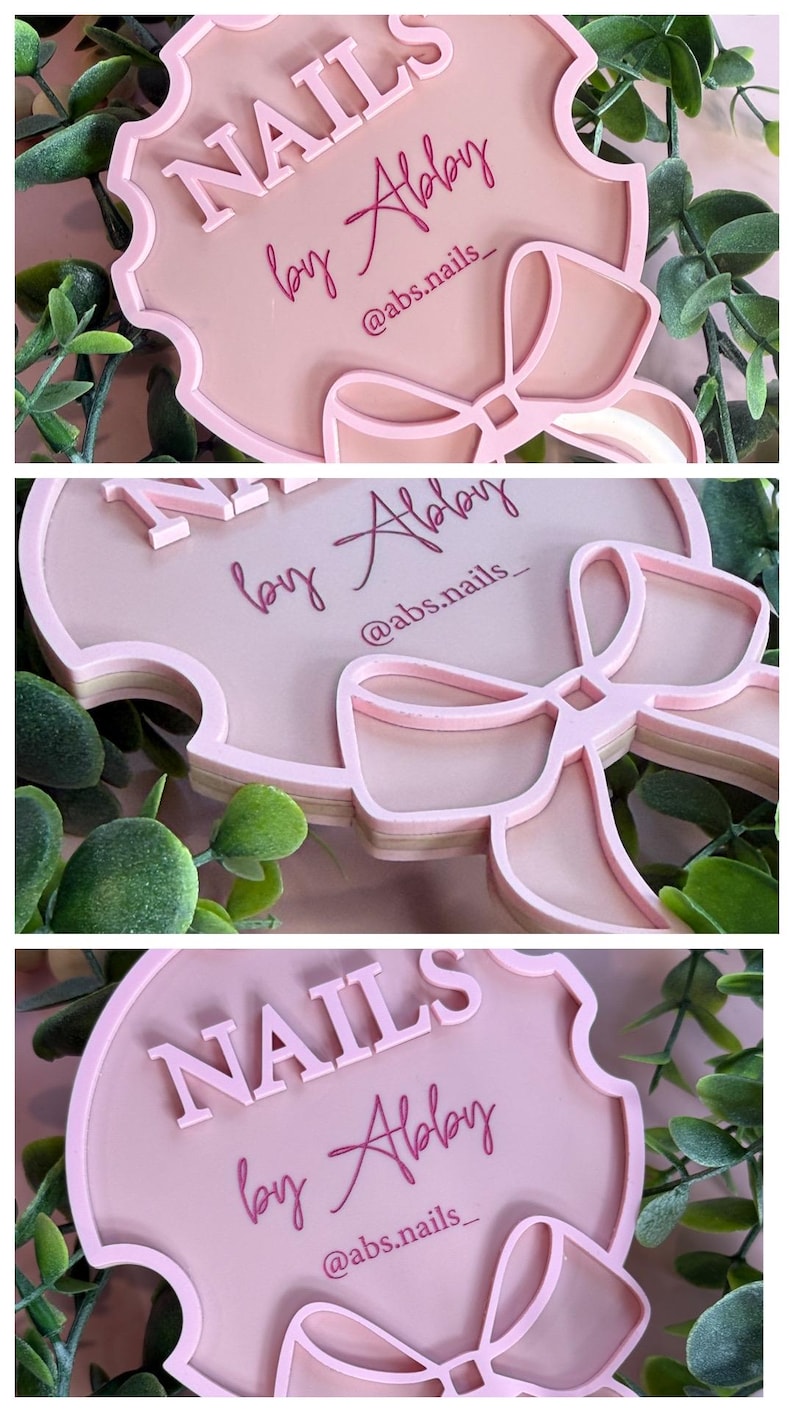 May include: A pink, layered sign with the text "NAILS by Abby" and the Instagram handle "@abs.nails_". The sign has a bow design at the bottom and is surrounded by green foliage.