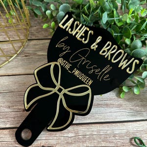 May include: A black handheld mirror featuring a gold bow graphic at the base and gold text that reads LASHES & BROWS by Griselle @GTHE_PMUQUEEN. The mirror has a circular cutout at the bottom of the handle and a smooth, reflective surface.