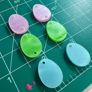 Könnte beinhalten: Sechs pastellfarbene Plastik-Ostereier mit einem Loch oben. Die Eier sind rosa, grün, hellblau und dunkelblau. Die Eier sind auf einer grünen Schneidematte angeordnet.