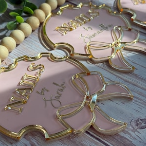 May include: Pink and gold decorative name tags with a bow design. The tags have a gold border and the name "NAILS" is displayed in gold lettering. The text "by Sarah" is also visible. The tags are arranged on a wooden surface.