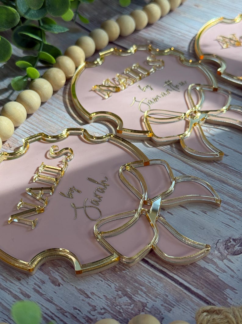 May include: Pink and gold decorative name tags with a bow design. The tags have a gold border and the name "NAILS" is displayed in gold lettering. The text "by Sarah" is also visible. The tags are arranged on a wooden surface.