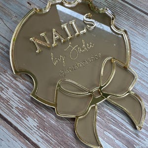 May include: A gold-colored acrylic sign with the text "NAILS by Jadee" in gold lettering. The sign has a decorative bow and a scalloped edge. The Instagram handle @setsbyssadee is also visible.