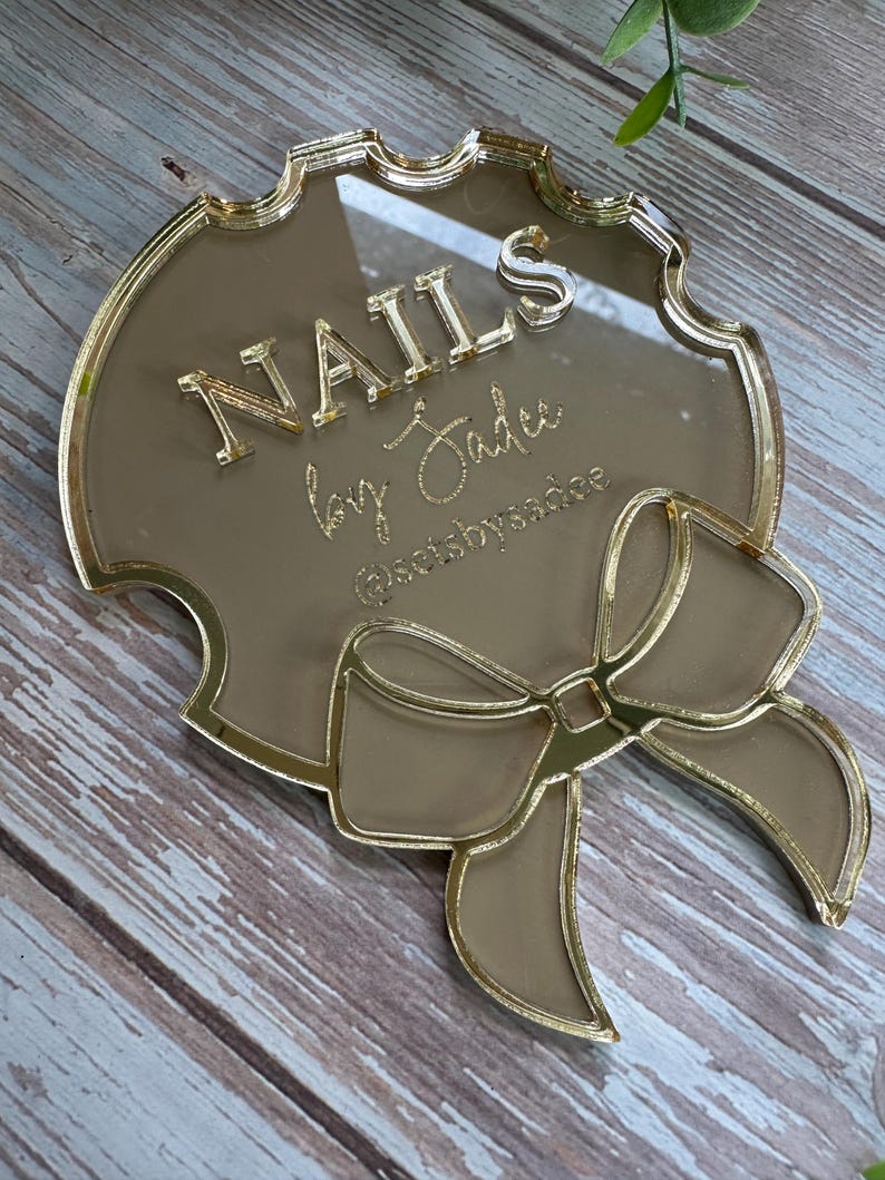 May include: A gold-colored acrylic sign with the text "NAILS by Jadee" in gold lettering. The sign has a decorative bow and a scalloped edge. The Instagram handle @setsbyssadee is also visible.