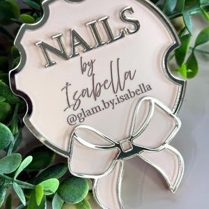 May include: A light pink sign with silver trim and a bow design. The sign reads "NAILS by Isabella" with the Instagram handle "@glam.by.isabella" below. The sign is surrounded by green foliage.