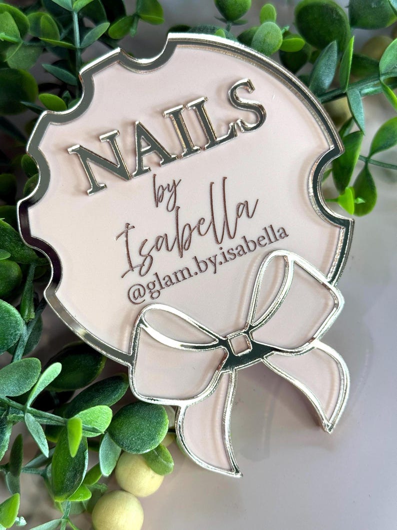 May include: A light pink sign with silver trim and a bow design. The sign reads "NAILS by Isabella" with the Instagram handle "@glam.by.isabella" below. The sign is surrounded by green foliage.