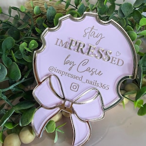 May include: A light pink, decorative plaque with gold trim and a bow design. The plaque features the text "stay IMPRESSED by Casie" and an Instagram handle. The plaque is surrounded by greenery.