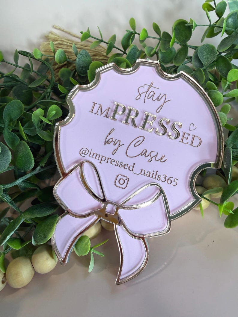 May include: A light pink, decorative plaque with gold trim and a bow design. The plaque features the text "stay IMPRESSED by Casie" and an Instagram handle. The plaque is surrounded by greenery.