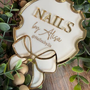 May include: A white and gold acrylic sign with the text "NAILS by Alisa" and the Instagram handle "@Nailzzby.lis". The sign has a bow design and is surrounded by greenery and wooden beads.