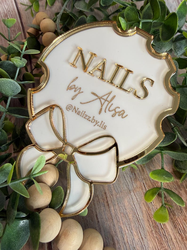 May include: A white and gold acrylic sign with the text "NAILS by Alisa" and the Instagram handle "@Nailzzby.lis". The sign has a bow design and is surrounded by greenery and wooden beads.