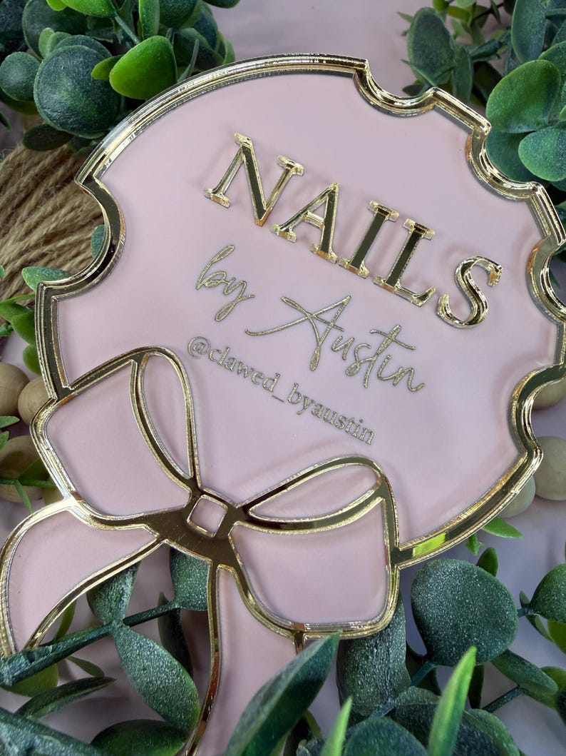 May include: A pink and gold decorative sign with the text "NAILS by Austin" and the Instagram handle "@clawed_byaustin". The sign has a bow design and a gold border.