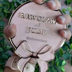 May include: A pink, round decorative item with gold trim and a bow design. The text "RAW CLAWS with JULIE" and the Instagram handle "@jujurawclaws" are displayed on the surface. The item is held against a backdrop of green foliage.
