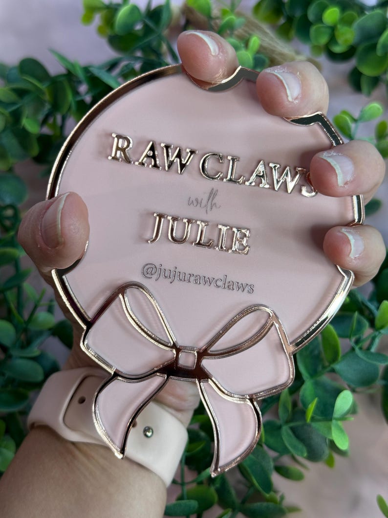 May include: A pink, round decorative item with gold trim and a bow design. The text "RAW CLAWS with JULIE" and the Instagram handle "@jujurawclaws" are displayed on the surface. The item is held against a backdrop of green foliage.
