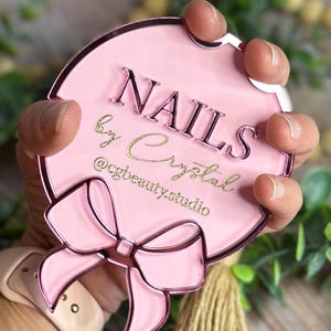 May include: A pink, round sign with the words "NAILS by Crystal" in a script font and the Instagram handle "@cgbeauty.studio". The sign has a bow design and a metallic pink border.