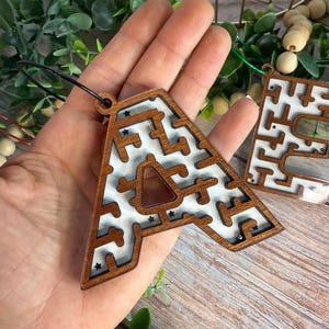 May include: A wooden letter A shaped maze puzzle with a white backing and a dark brown frame. The maze features small black star accents and a metal ball bearing inside the path. A black cord is attached to the top for hanging as an ornament.