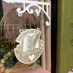Pode incluir: Uma placa branca e dourada com a palavra "ROMANCE" acima de um livro aberto, uma pena e um design de coração. A placa está pendurada em um suporte branco decorativo. A placa está em frente a uma coleção de livros.