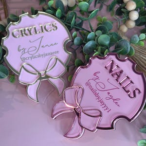 May include: Two pink and gold acrylic signs with bow accents. One sign reads "CRYILCS by Janae @crylicsbyjanaeee", the other reads "NAILS by Jaydee @nailsby.jayyy1". The signs are surrounded by greenery.
